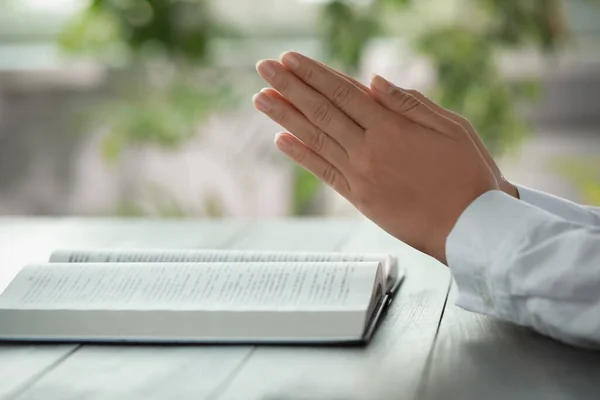 Woman holding hands clasped while praying at grey wooden table with Bible, closeup