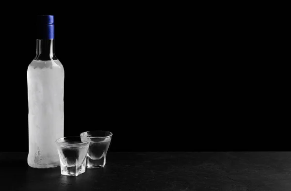 Bottle of vodka and shot glasses on table against black background. Space for text