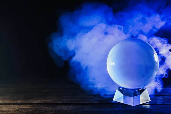 Magic crystal ball on wooden table and smoke against dark background. Making predictions