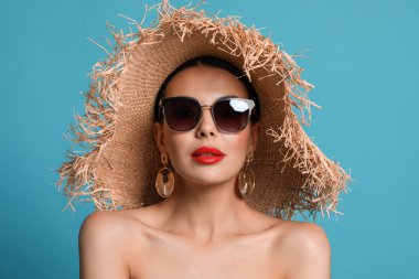Modaya uygun güneş gözlüklü ve açık mavi arka planda hasır şapkalı çekici bir kadın.