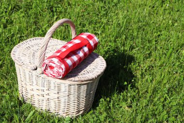 Rolled checkered tablecloth with picnic basket on green grass outdoors, space for text