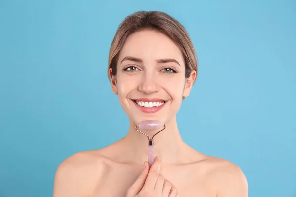 Young woman using natural rose quartz face roller on light blue background