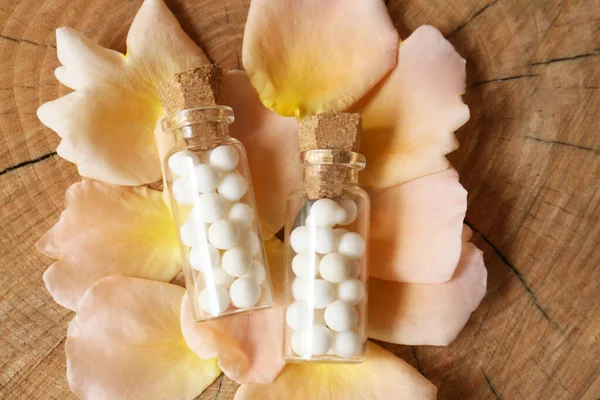 Bottles of homeopathic remedy and flower petals on wooden background, flat lay