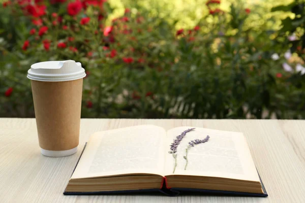 Open book with paper cup of coffee and dried flowers on wooden table in garden