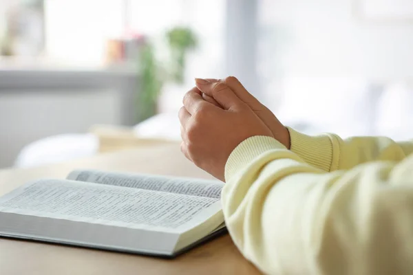 Woman holding hands clasped while praying at wooden table with Bible indoors, closeup. Space for text