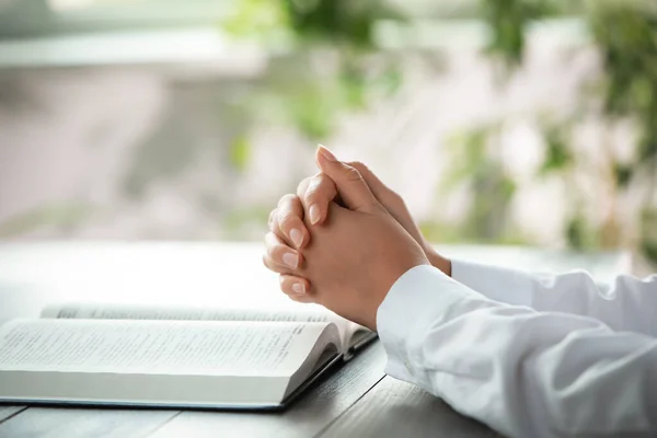 Woman holding hands clasped while praying at grey wooden table with Bible, closeup