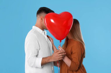 Açık mavi arka planda kalp şeklinde bir balonun arkasında öpüşen sevimli bir çift. Sevgililer Günü kutlaması