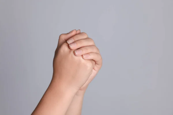 Woman holding hands clasped while praying against light grey background, closeup. Space for text