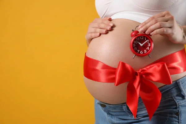 目覚まし時計と黄色の背景に赤い弓を持つ若い妊婦 クローズアップ 出産までの時間 — ストック写真