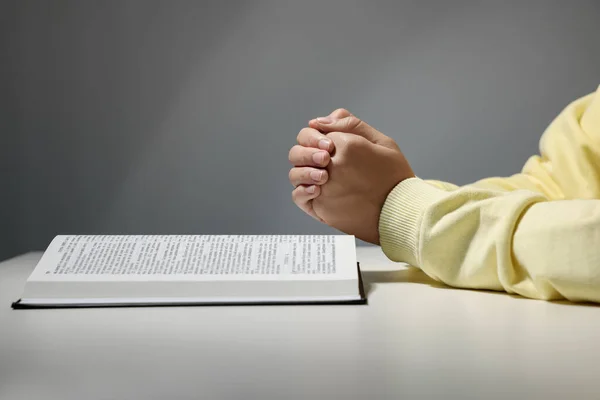 Woman praying over Bible at white table against grey background, closeup