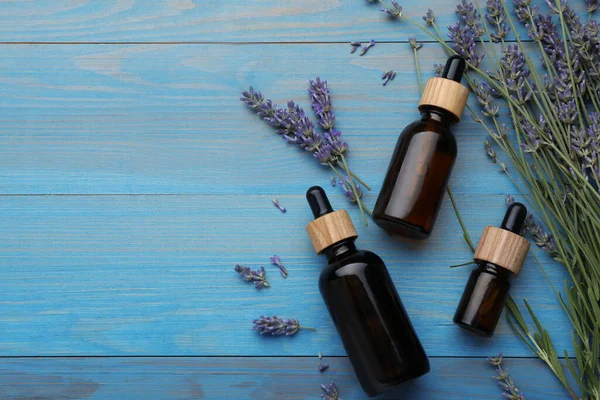 Bottles of essential oil and lavender flowers on light blue wooden table, flat lay. Space for text