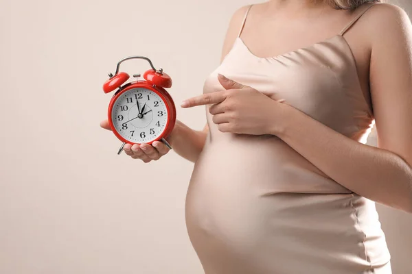 ベージュを背景に腹近くの目覚まし時計を指差す若い妊婦さんが近づいています 出産までの時間 — ストック写真