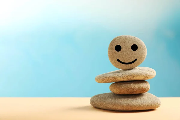 Stack of stones with drawn happy face on beige table against light blue background, space for text. Zen concept