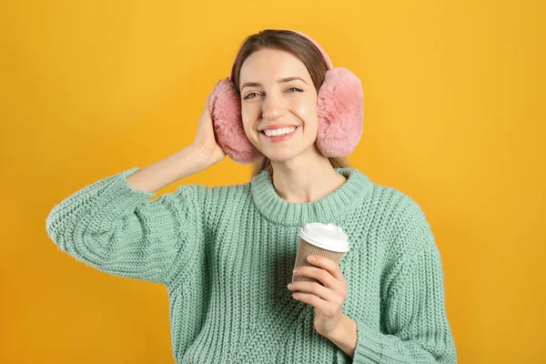 黄色の背景に暖かい耳飾りを身に着けている飲み物のカップと幸せな女性 — ストック写真