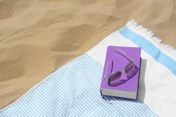 Beach towel with book and sunglasses on sand, space for text