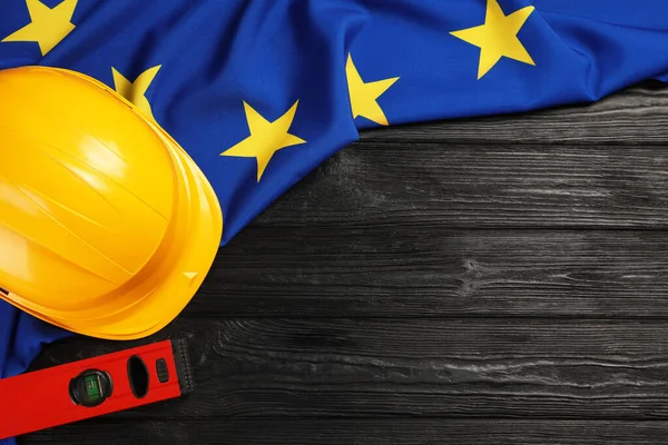 Yellow hard hat, building level and flag of European Union on black wooden table, flat lay. Space for text