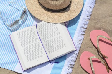 Beach towel with book, straw hat, sunglasses and flip flops on sand, flat lay