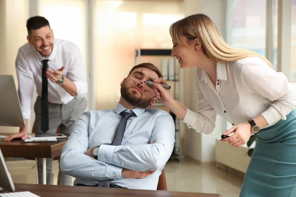 Young woman drawing on colleague\'s face while he sleeping in office. Funny joke
