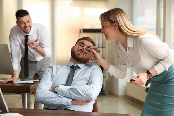 Young woman drawing on colleague's face while he sleeping in office. Funny joke