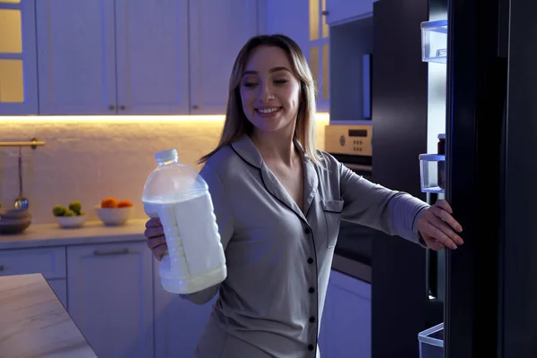 Young woman holding gallon bottle of milk near refrigerator in kitchen at night