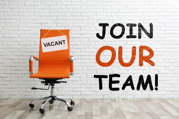 Join our team! Orange office chair with sign VACANT near white brick wall indoors