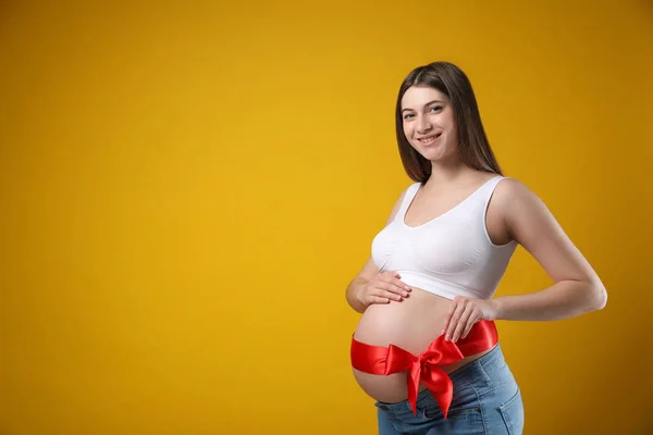 黄色の背景 テキストのためのスペースに対して彼女の腹の上に赤い弓を持つ若い妊婦 出産までの時間 — ストック写真