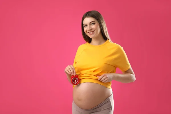 ピンクの背景に腹の近くに目覚まし時計を持っている若い妊婦 出産までの時間 — ストック写真