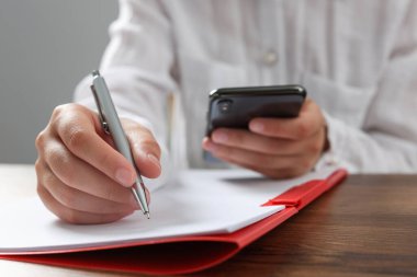 Ofisteki ahşap masadaki kırmızı klasörde akıllı telefon kağıtlarına yazı yazan kadın.