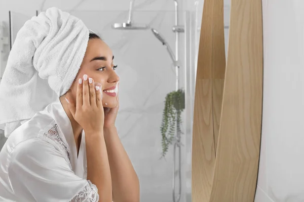 バスルーム テキストのためのスペースでミラーの近くに顔にクレンジングフォームを適用する美しい若い女性 スキンケア化粧品 — ストック写真