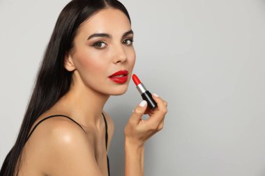 Gri arkaplanda kırmızı ruj tutan güzel makyajlı genç bir kadın.
