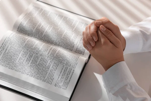 Woman holding hands clasped while praying over Bible at white table, closeup