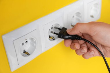 Woman inserting plug into power socket on yellow wall, closeup. Electrical supply