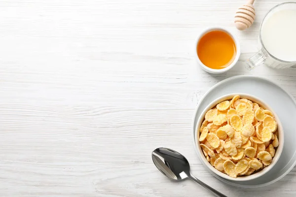 Tasty crispy corn flakes served with milk and honey on white wooden table, flat lay. Space for text