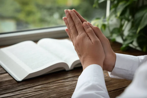 Woman holding hands clasped while praying at wooden table with Bible, closeup. Space for text