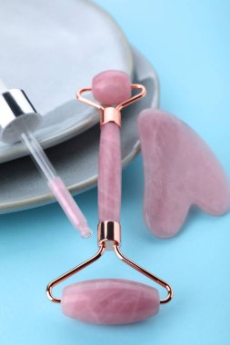 Gül kuvars gua sha aracı, yüz silindiri ve açık mavi arka planda damlatıcı, yakın plan