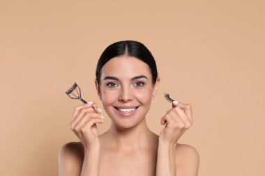 Beautiful young woman with false eyelashes and curler on beige background