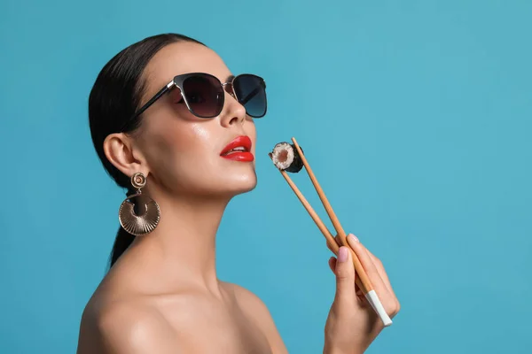 Attractive woman in fashionable sunglasses holding chopsticks with sushi against light blue background