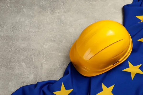 Yellow hard hat and flag of European Union on light grey table, above view. Space for text