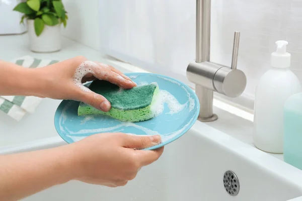Woman washing plate with sponge above sink in kitchen, closeup