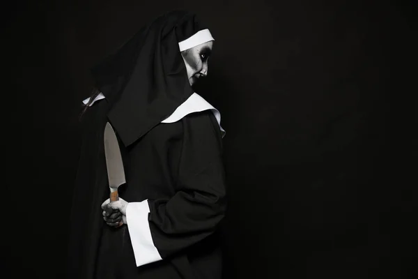 Scary devilish nun with knife behind back on black background, space for text. Halloween party look