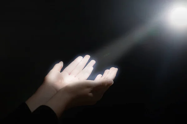 Woman stretching hands towards light in darkness, closeup. Praying concept