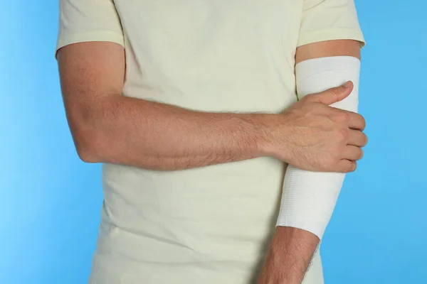 Man with arm wrapped in medical bandage on light blue background, closeup