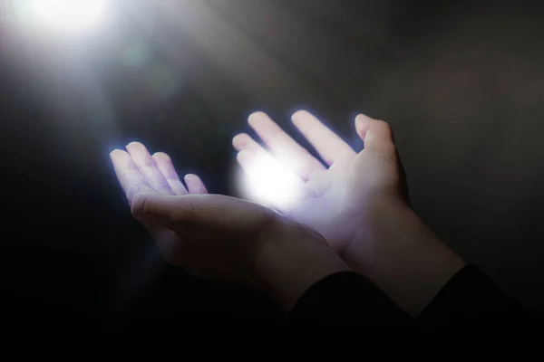 Woman stretching hands towards light in darkness, closeup. Praying concept