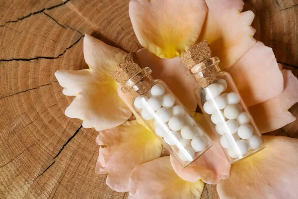 Bottles of homeopathic remedy and flower petals on wooden background, flat lay