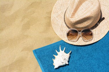 Yumuşak mavi plaj havlusu, güneş gözlüğü, hasır şapka ve kumdaki deniz kabuğu. Metin için boşluk