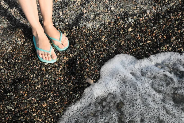 Woman in stylish flip flops on pebble beach near beautiful sea wave, closeup