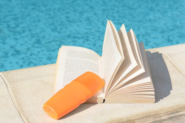 Open book and sunscreen on swimming pool edge