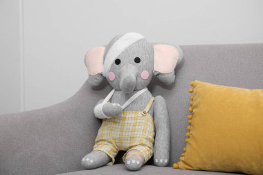 Bandajlı oyuncak fil kanepede, ışık duvarının yanında oturuyor.