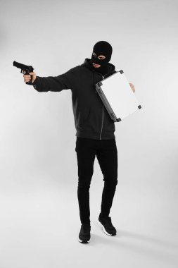 Metal çantalı, siyah kar maskeli ve açık gri arka planda silahlı bir adam.