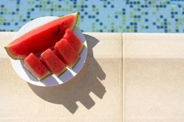 Slices of watermelon on white plate near swimming pool outdoors, top view. Space for text